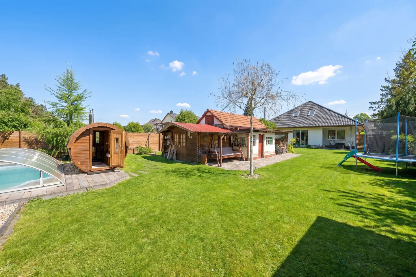 A lush green backyard with a pool, sauna, shed, swing, trampoline, and slide under a bright blue sky.