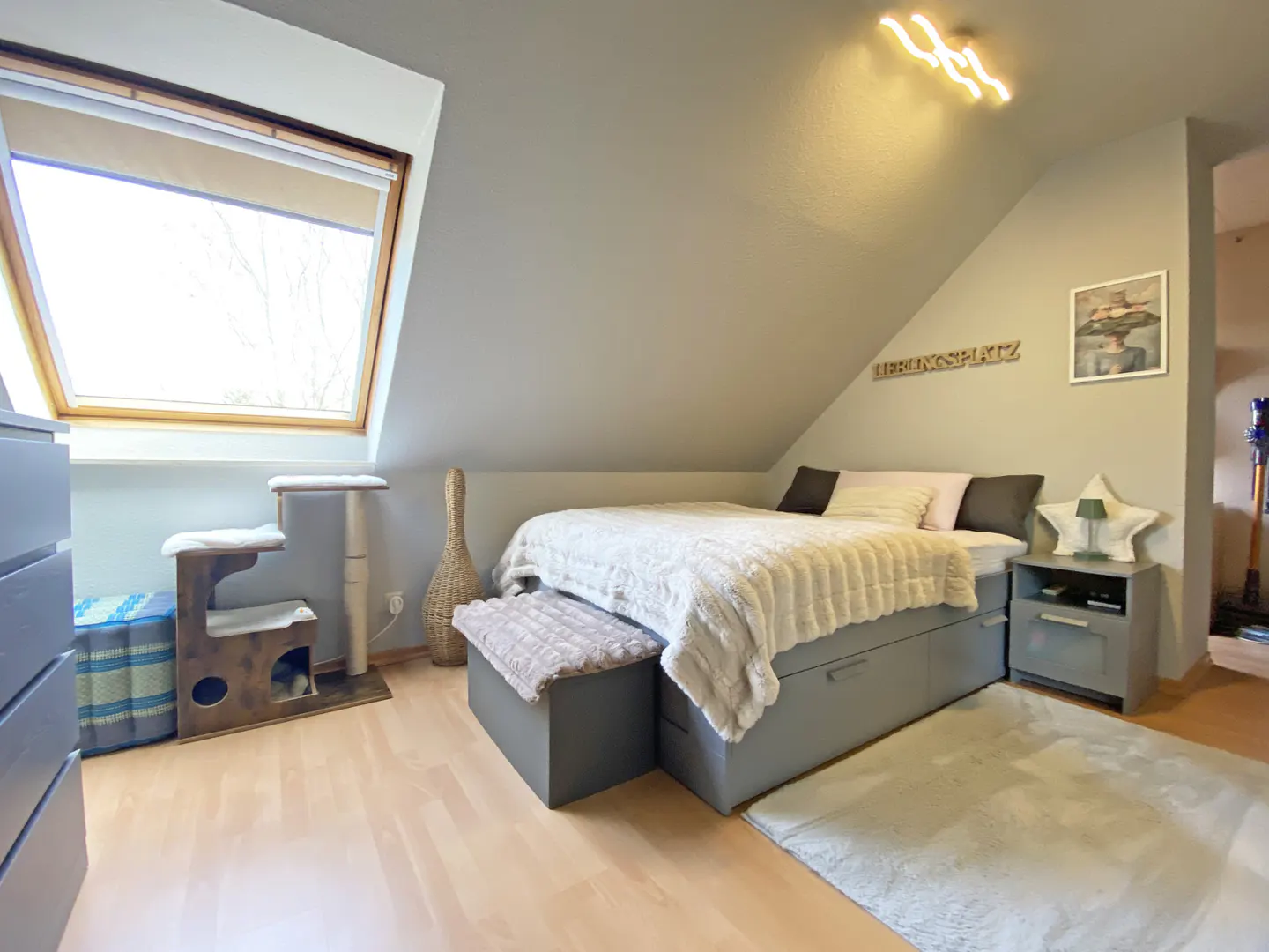 Attic bedroom with gray walls, wood floors, and a large window. A bed with a white comforter and gray frame sits under the sloped ceiling. A cat tree is near the window.