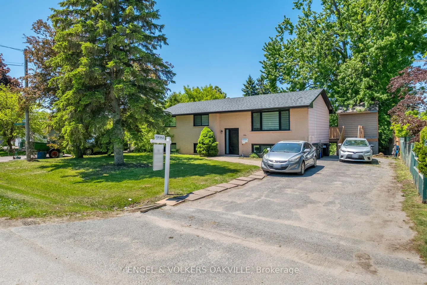 Single-story beige house with a dark roof, large front lawn, and a paved driveway with two parked cars on a sunny day.
