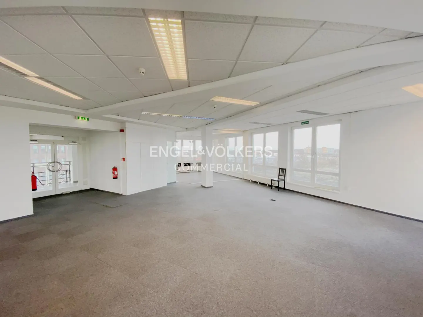 Bright, empty office space with white walls, gray floors, and fluorescent lighting. Windows line one wall, and a black chair sits near them. Fire extinguishers are visible near the entrance.