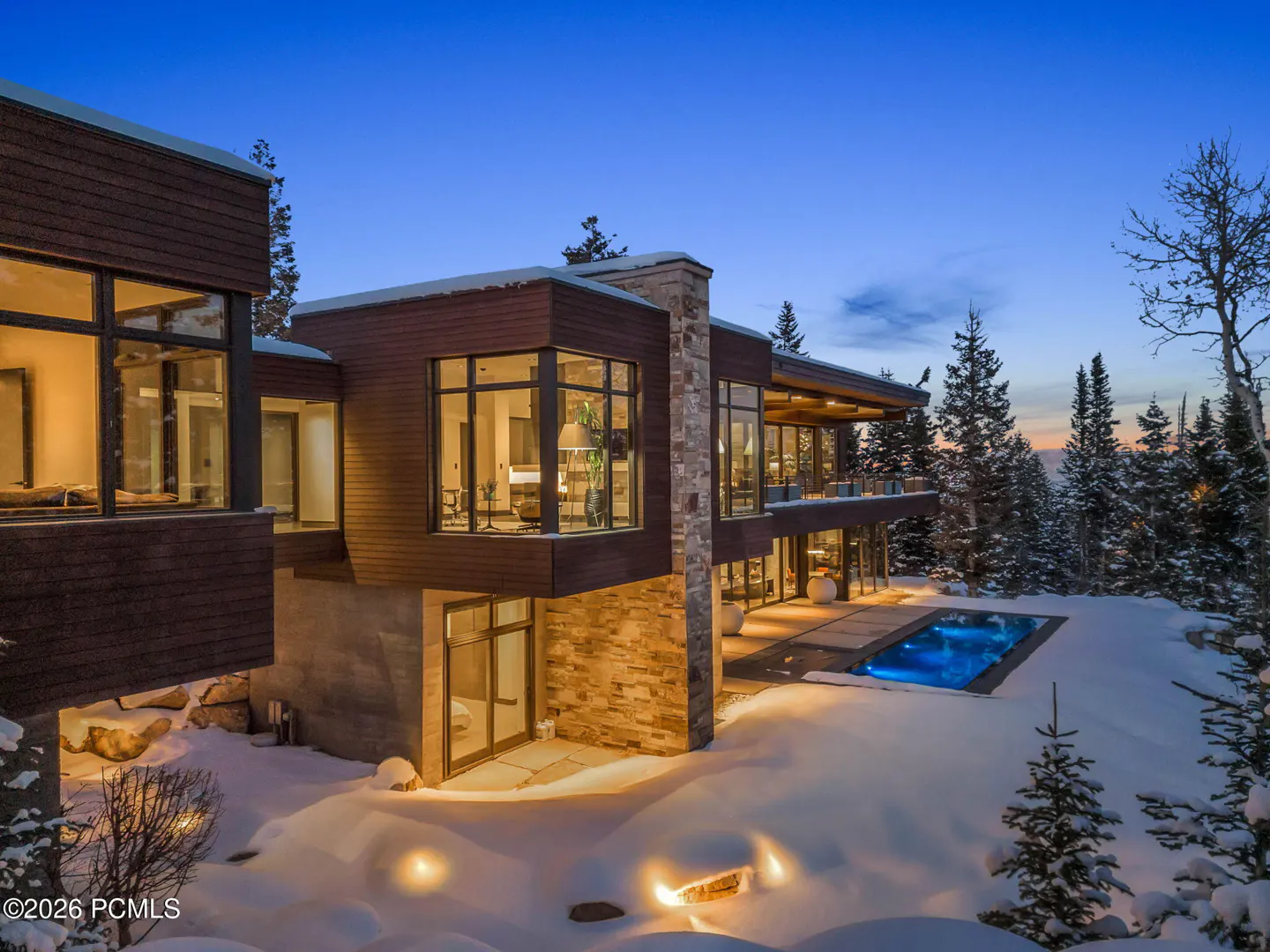 Modern mountain home with wood and stone exterior, illuminated pool, and snow-covered landscape at twilight.