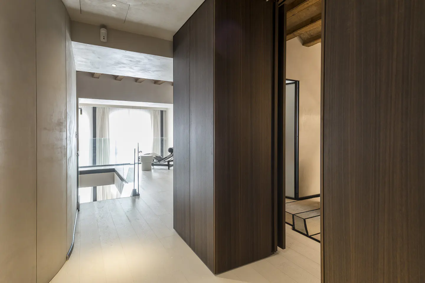 Hallway with dark wood sliding doors, light wood floors, and a view of a lounge chair by a window.