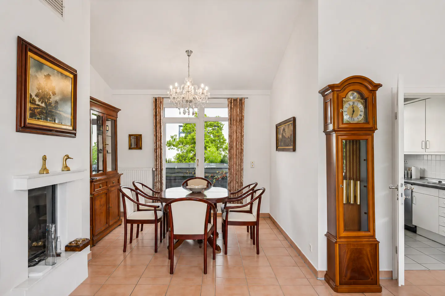 Dining room with a wooden table, six chairs, a crystal chandelier, a grandfather clock, and a fireplace on tiled floors.