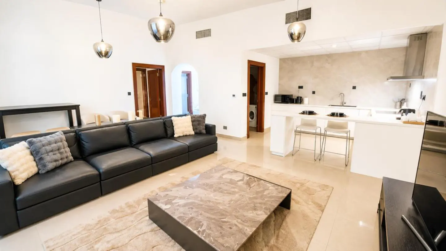 A modern apartment interior with a black leather sofa, marble coffee table, and open kitchen with white countertops.