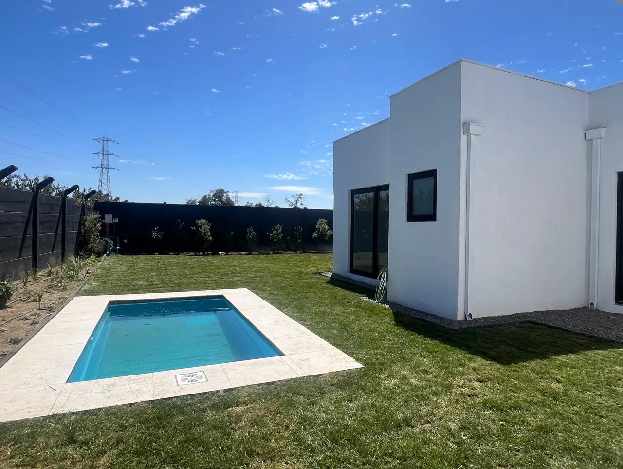 Backyard view of a modern white house with a small, rectangular pool surrounded by green grass and a dark fence. Blue sky overhead.