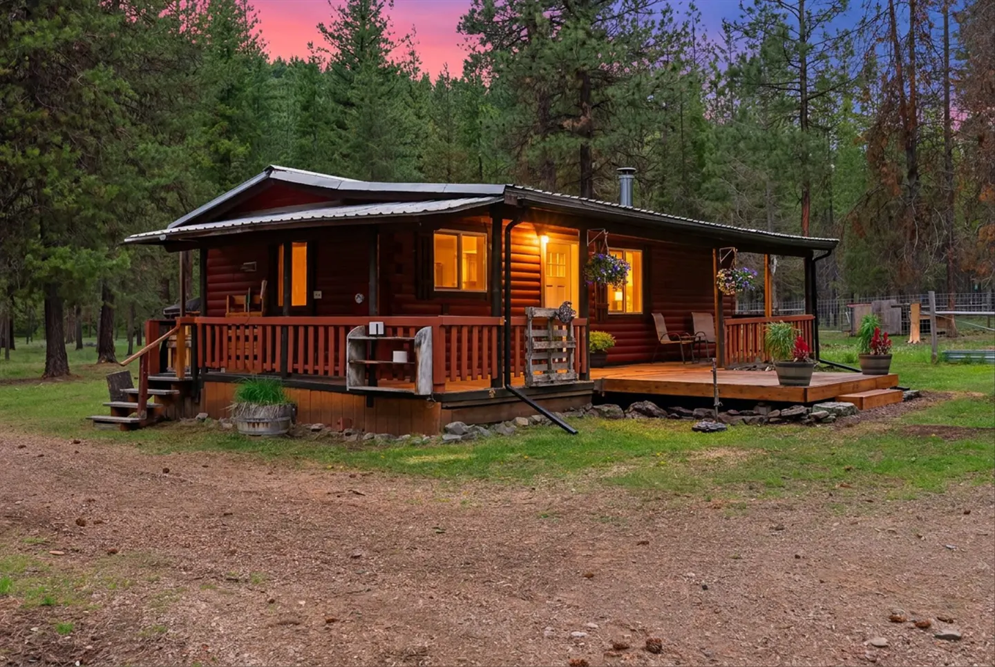 A cozy, red log cabin with a porch, surrounded by tall green trees under a colorful sunset sky.