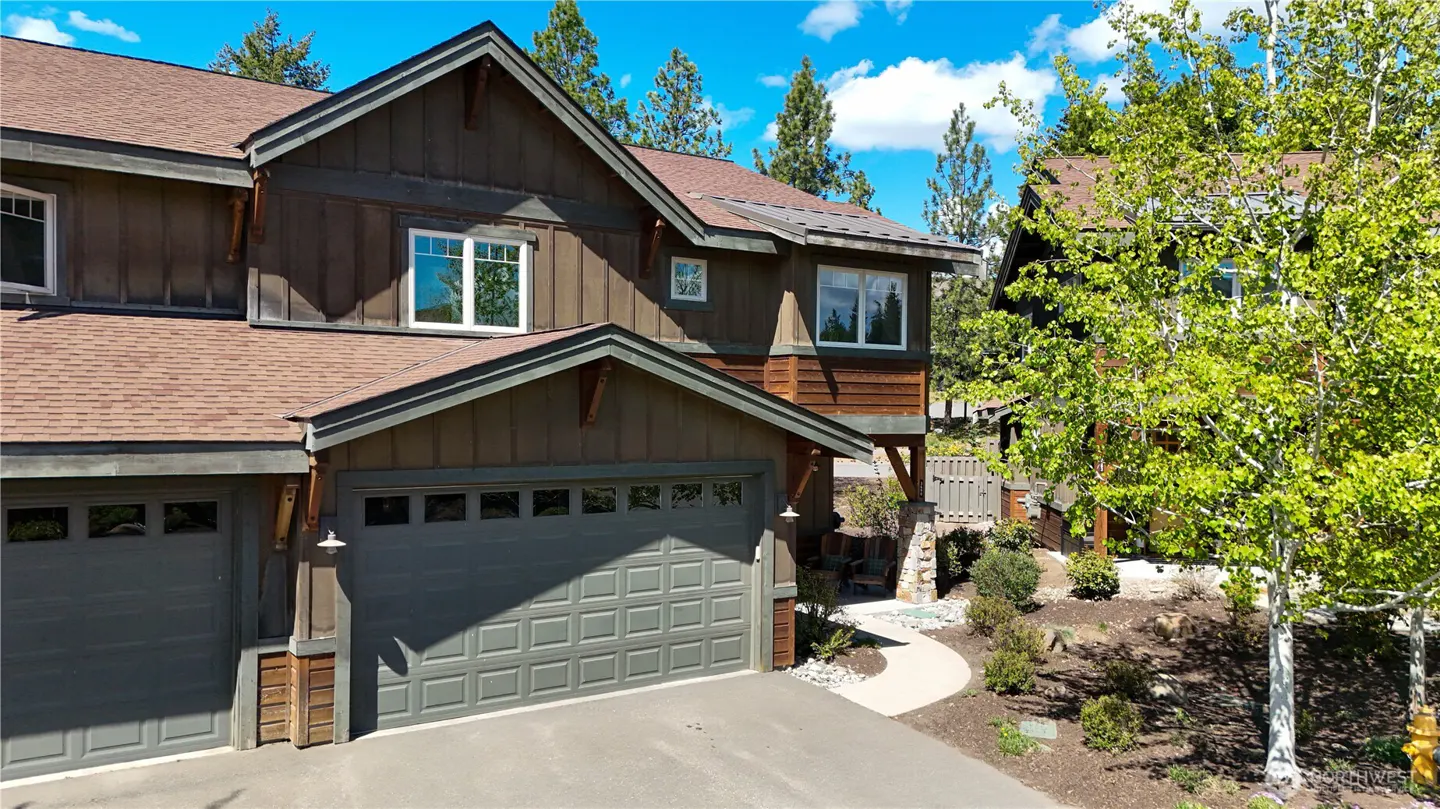Two-story brown wood-sided home with a gray garage, asphalt driveway, and green trees under a bright blue sky.