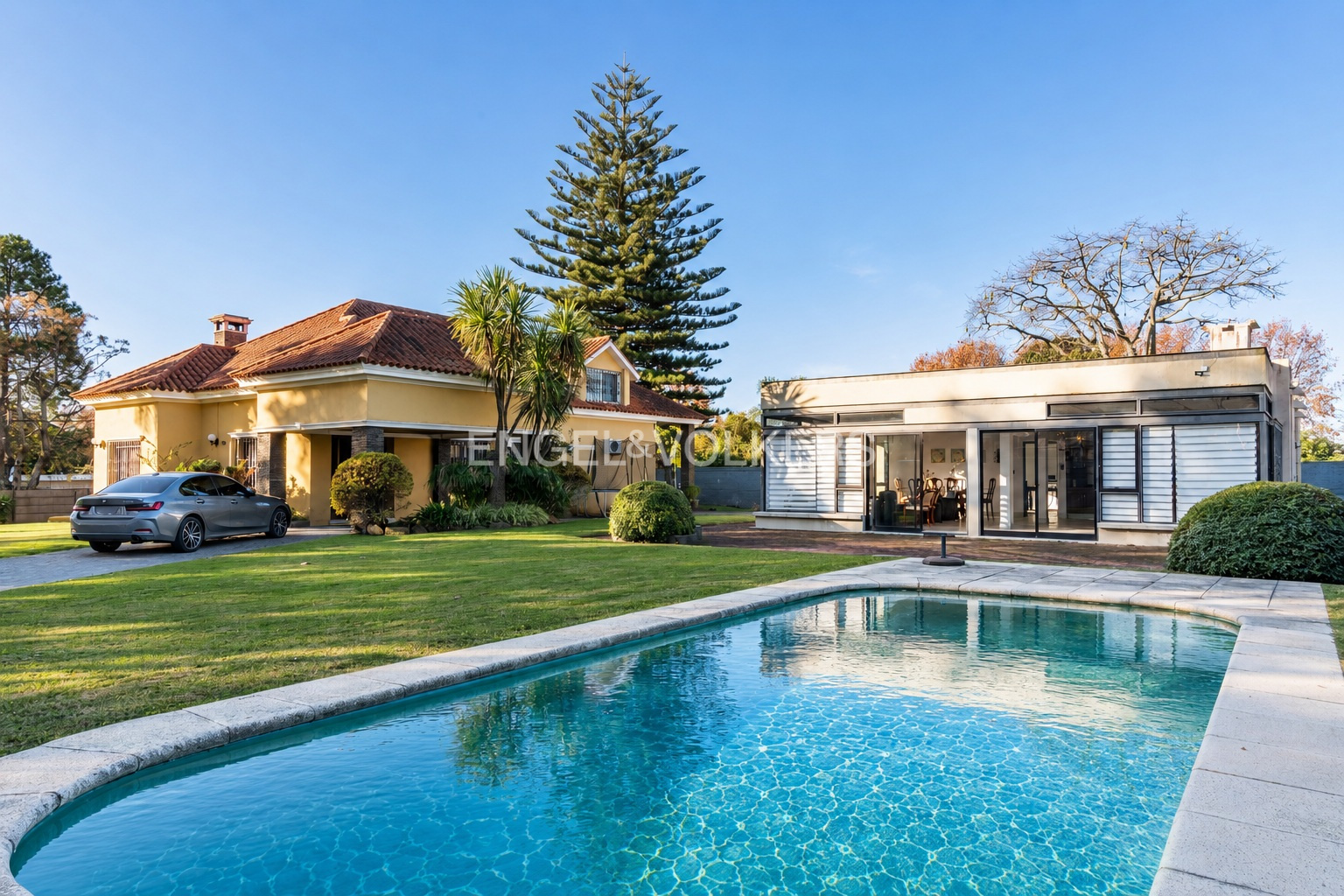 A yellow house and a modern glass-walled structure sit behind a swimming pool on a sunny day with a large pine tree.