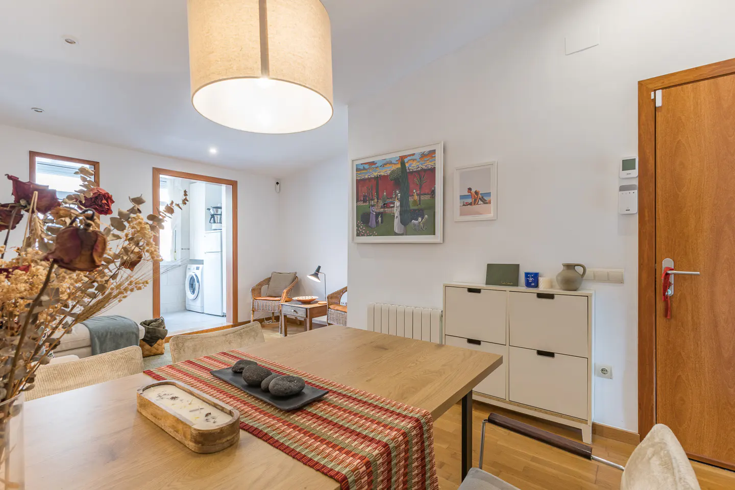 A bright living room with a wooden table, a striped runner, and decorative stones. White walls, artwork, and a glimpse into the kitchen.
