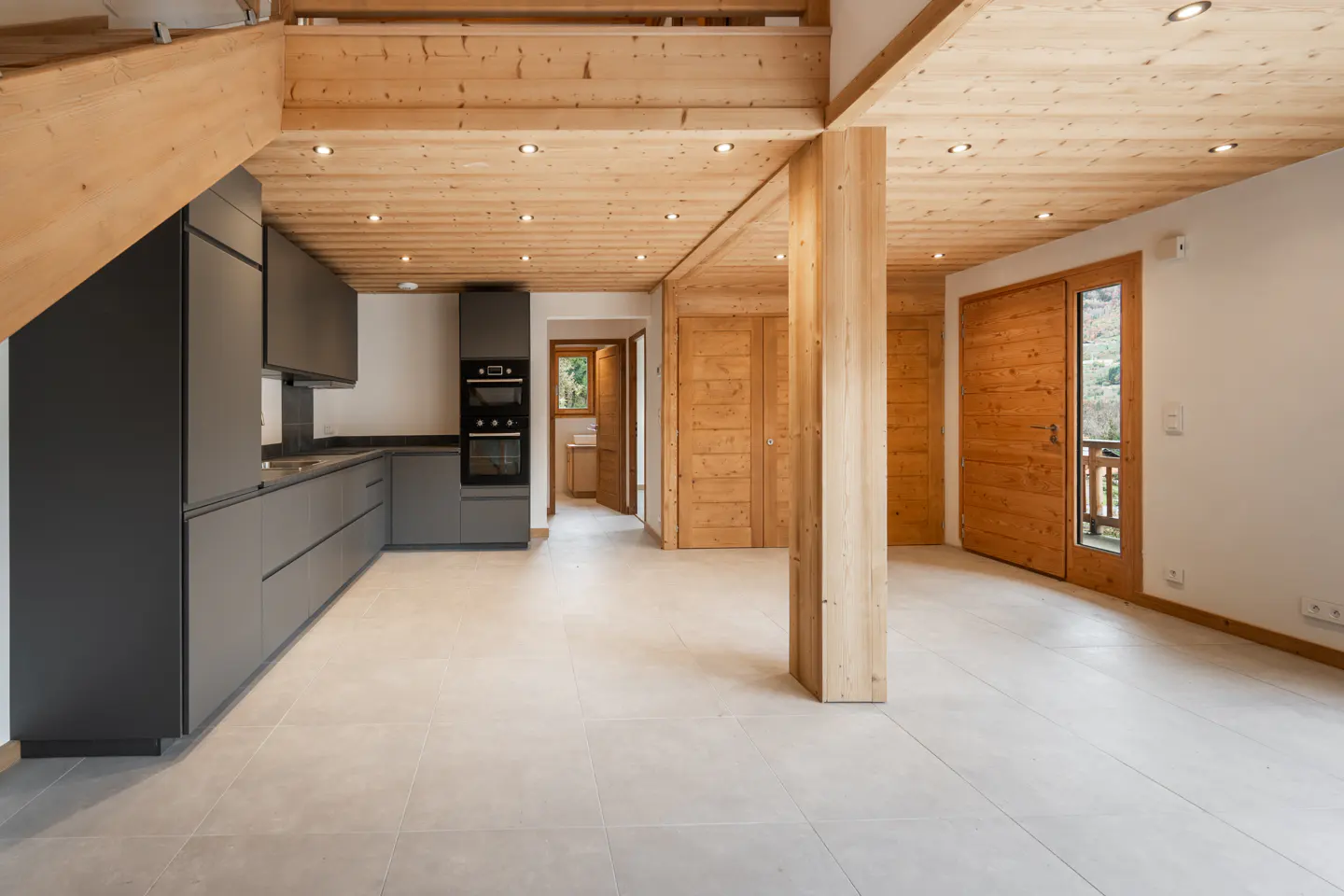 A modern kitchen and living area with gray cabinets, wood beams, and light tile flooring.