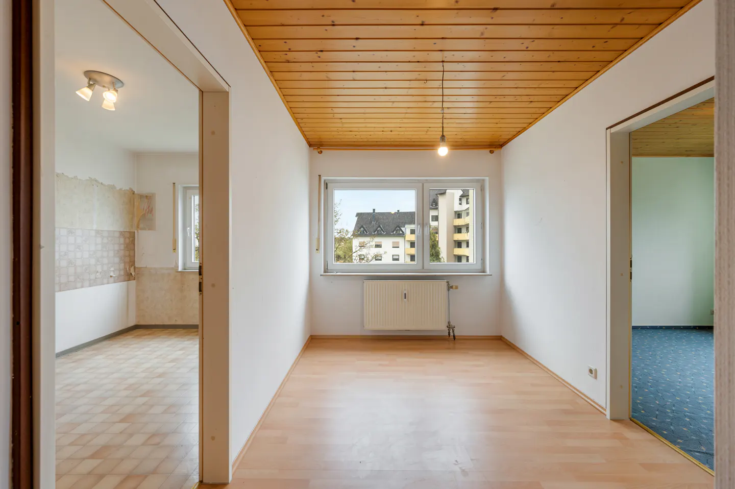 Empty room with wood-paneled ceiling, white walls, and wood floor. A window looks out to a building. Doorways lead to other rooms.