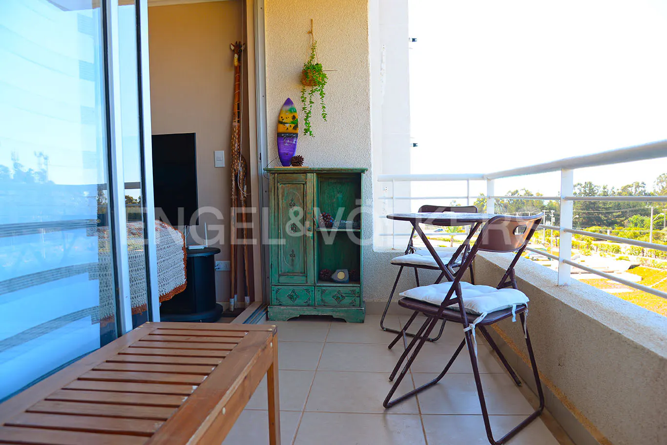 Balcony view with a wooden bench, table and chairs, a green cabinet, and a view of trees in the background.