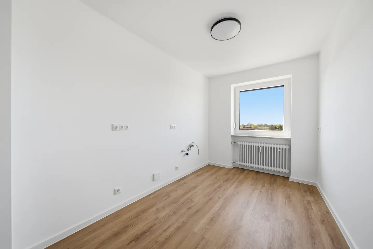 Bright, empty room with wood floor, white walls, and a window showing a blue sky. A radiator sits under the window.