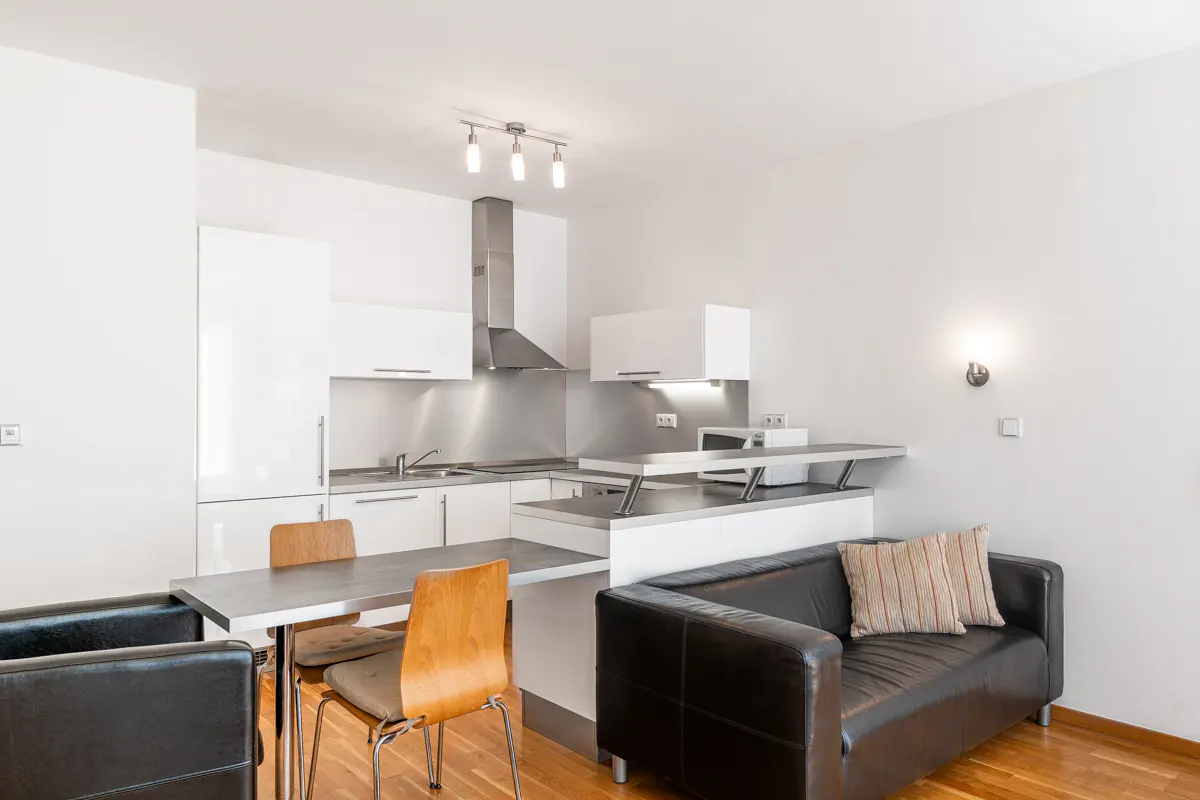 Bright, modern apartment interior with white walls, wood floors, and a black leather sofa. A kitchen with stainless steel appliances is visible.