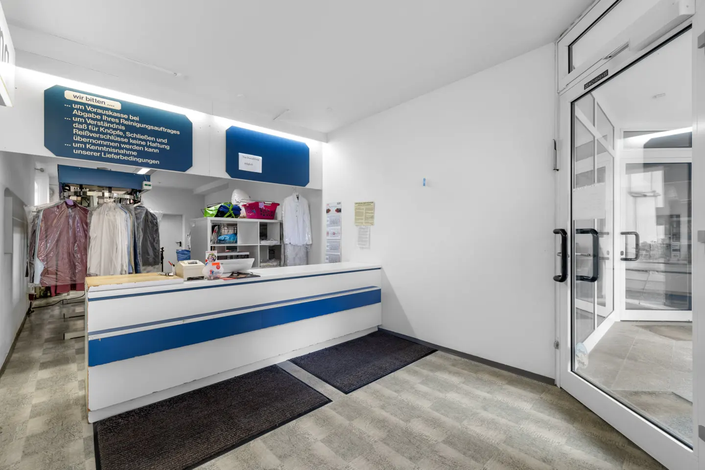 Interior of a dry cleaner with a white and blue service counter, hanging garments, and a glass entrance door.
