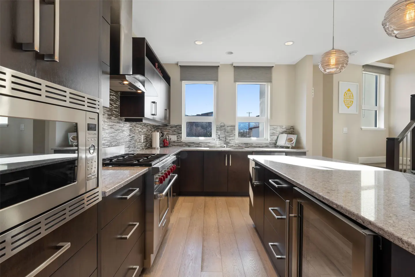 A modern kitchen with dark wood cabinets, stainless steel appliances, granite countertops, and light wood floors. Two windows offer a view of the outdoors.