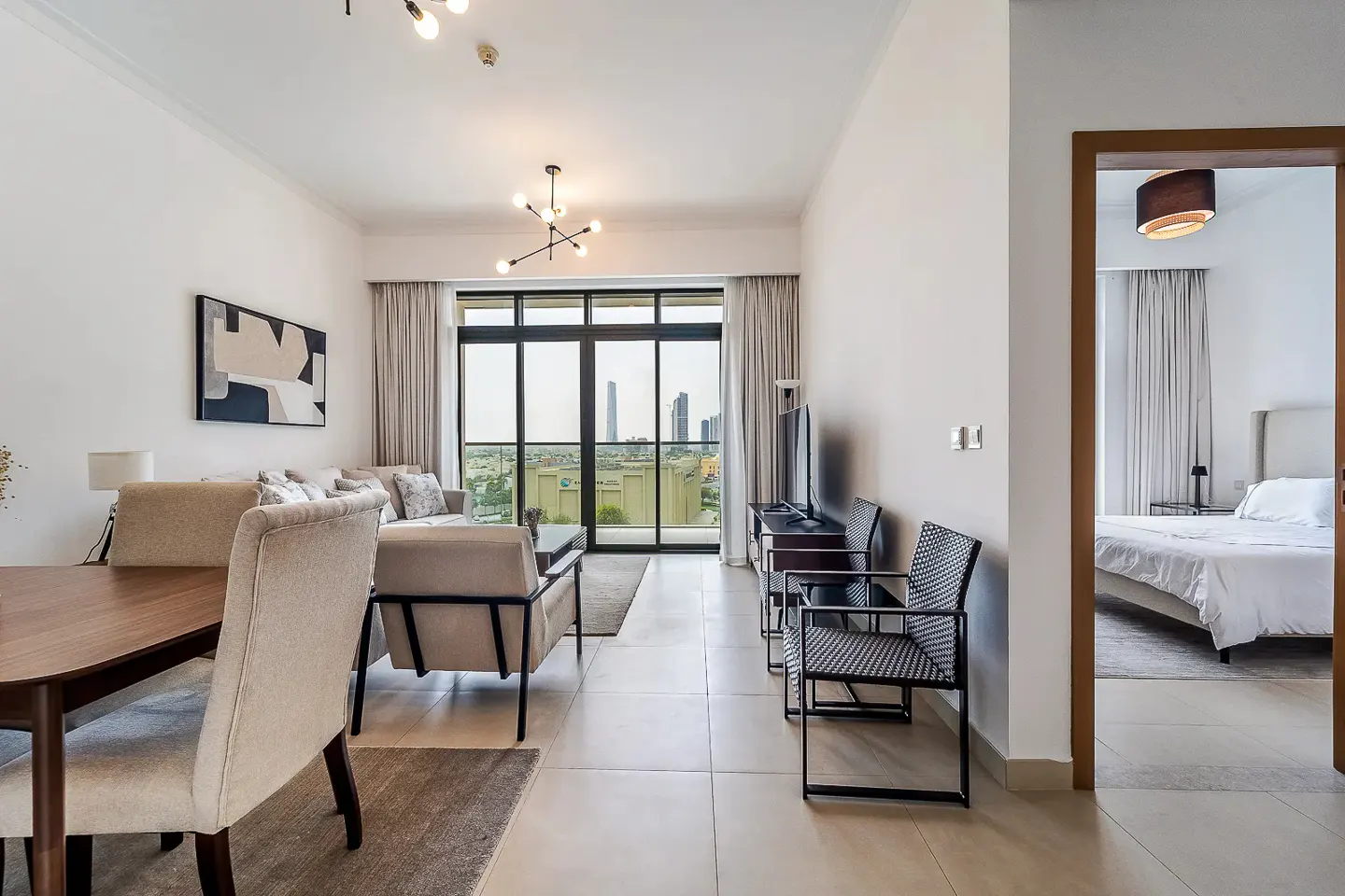 Bright, modern apartment interior with a dining table, sofa, and balcony view of city buildings. A bedroom is visible through a doorway.