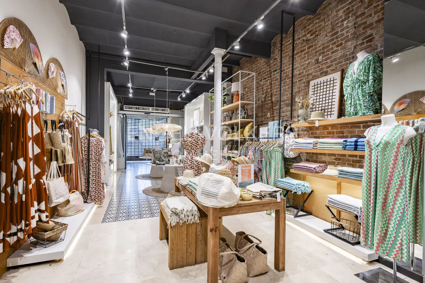 Retail store interior with clothing racks, wooden tables, and exposed brick walls. Mannequins display colorful dresses. Natural light illuminates the space.