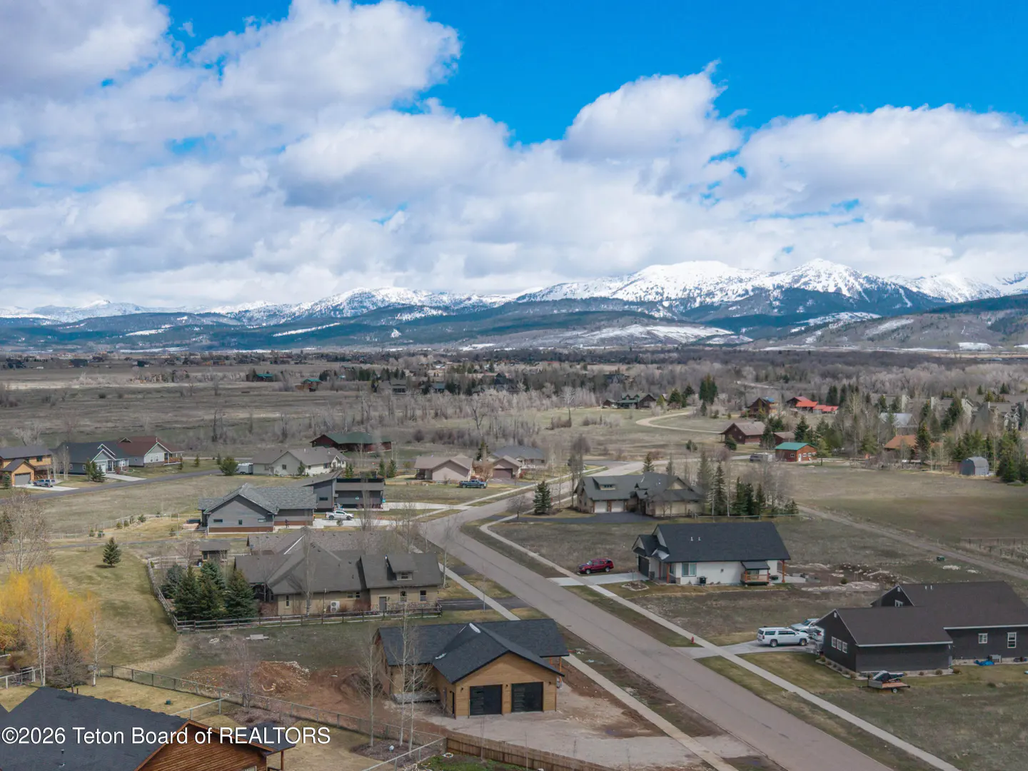 Aerial view of a residential area with houses, roads, and snow-capped mountains in the background under a blue sky with white clouds.