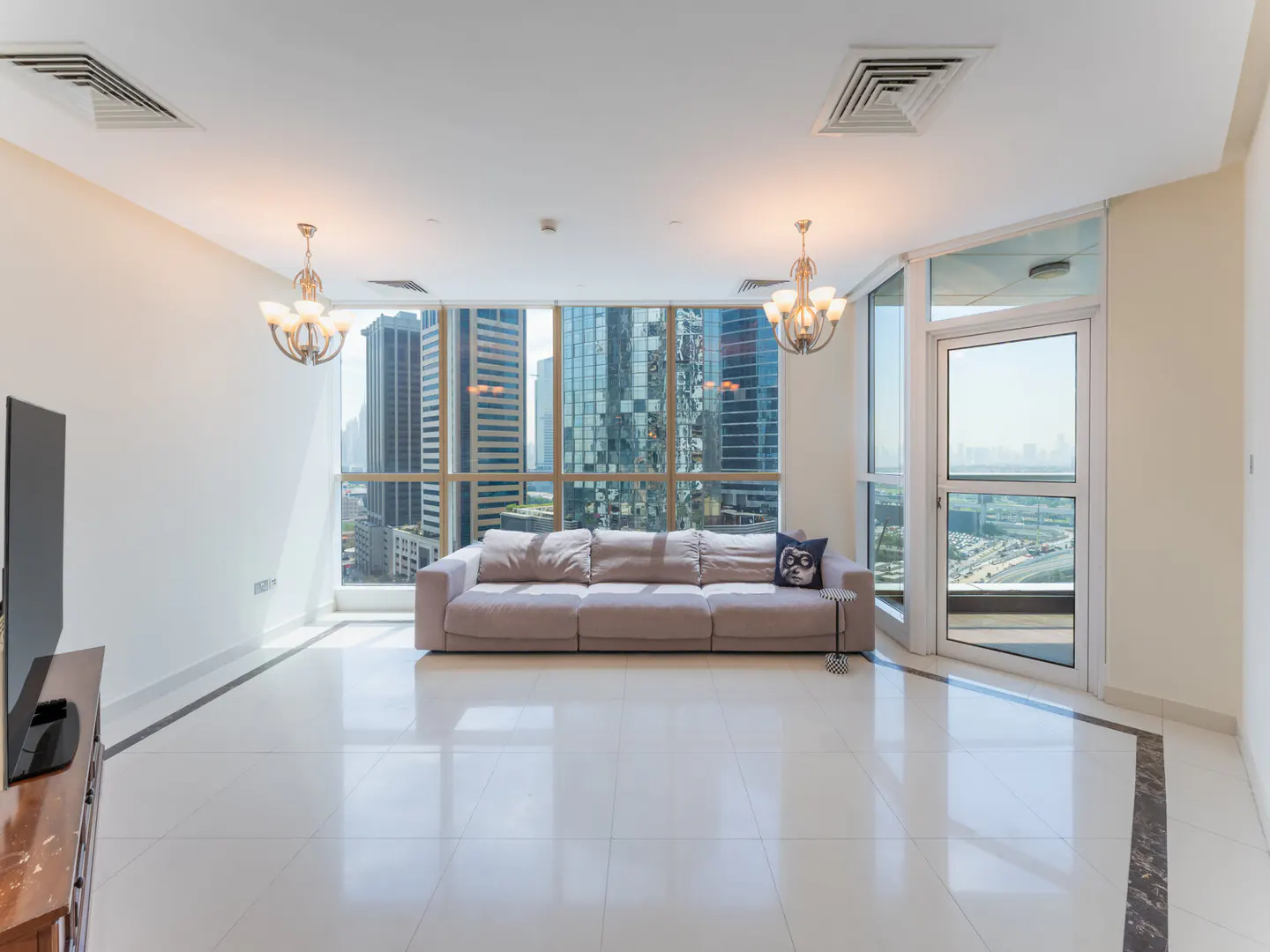 Bright, modern living room with a long gray sofa facing large windows with a city view. Chandeliers hang from the ceiling.