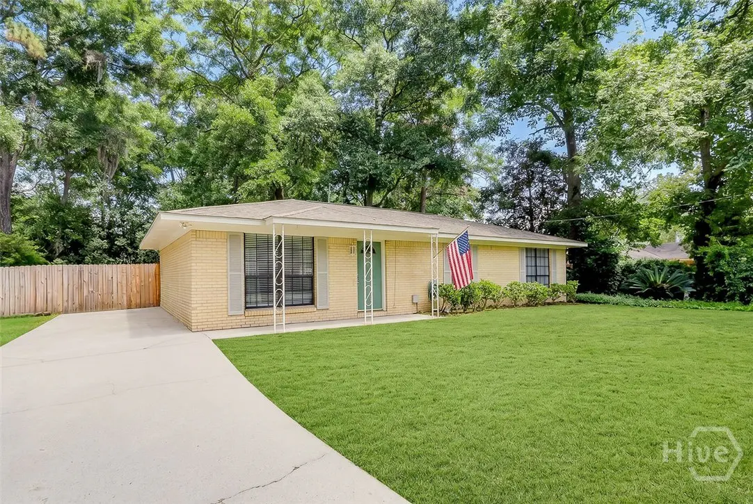 A single-story, light-yellow brick house with a green door, gray shutters, and an American flag on a green lawn.