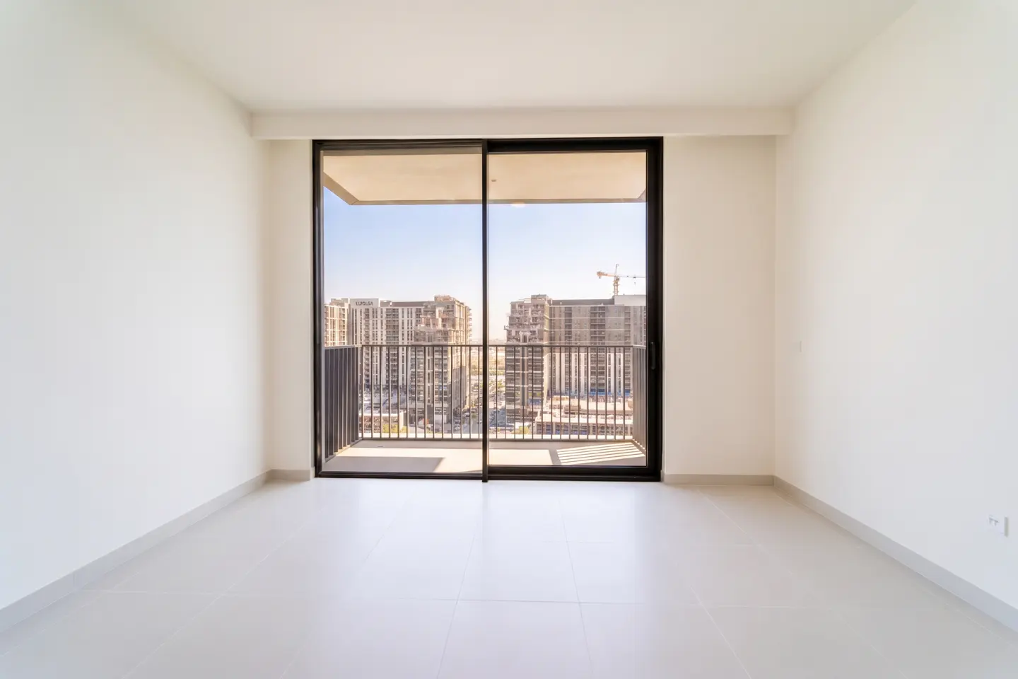 Empty white room with sliding glass doors to a balcony overlooking city buildings under a blue sky.