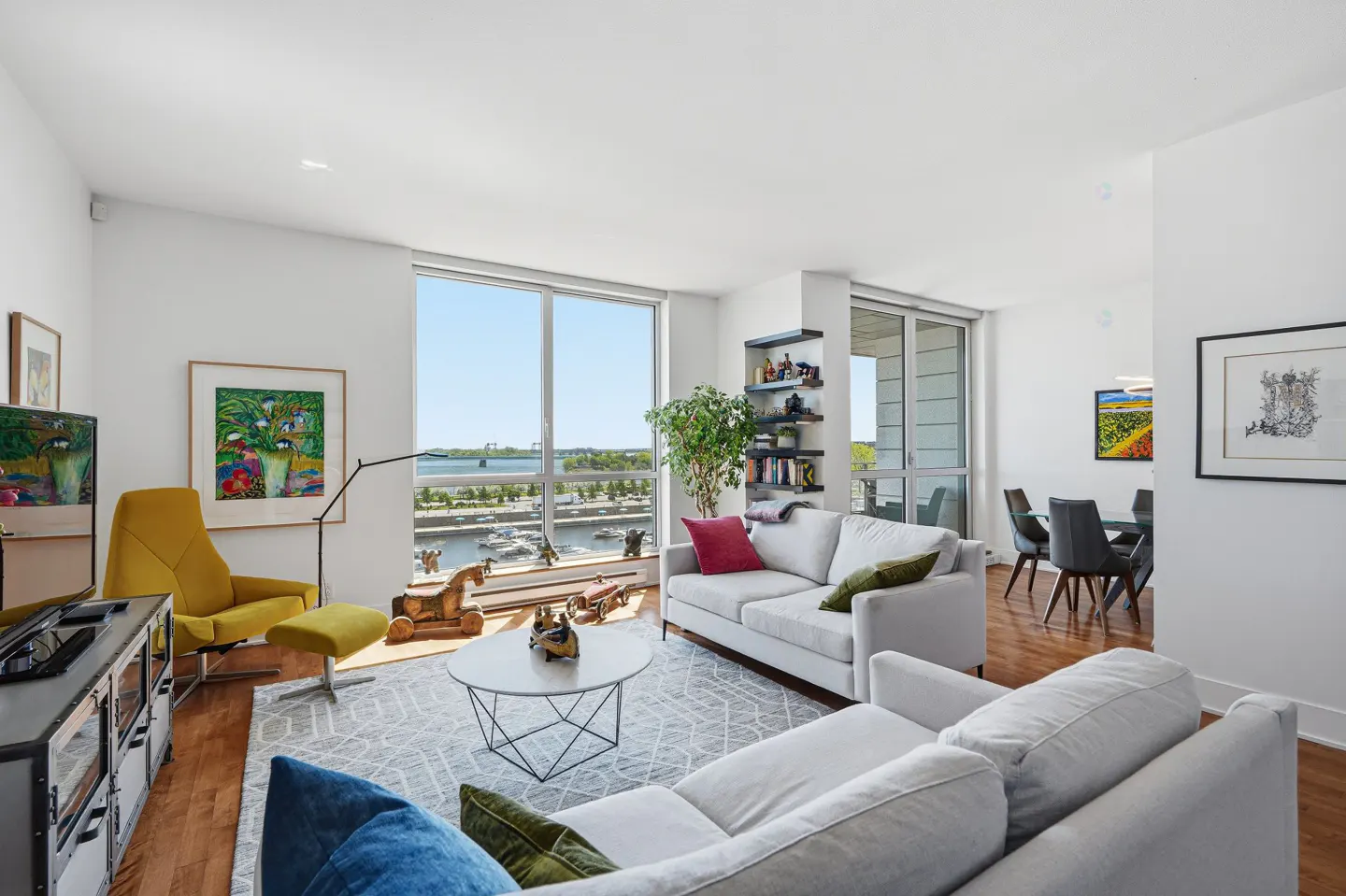 Bright, modern living room with white walls, gray sofas, yellow chair, and a view of a river and boats.