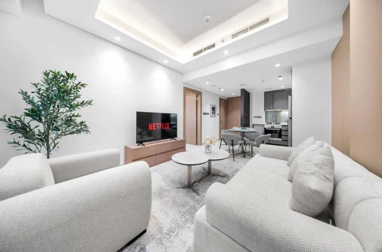 Bright living room with white sofas, a TV showing Netflix, and a small dining area leading to a gray kitchen.