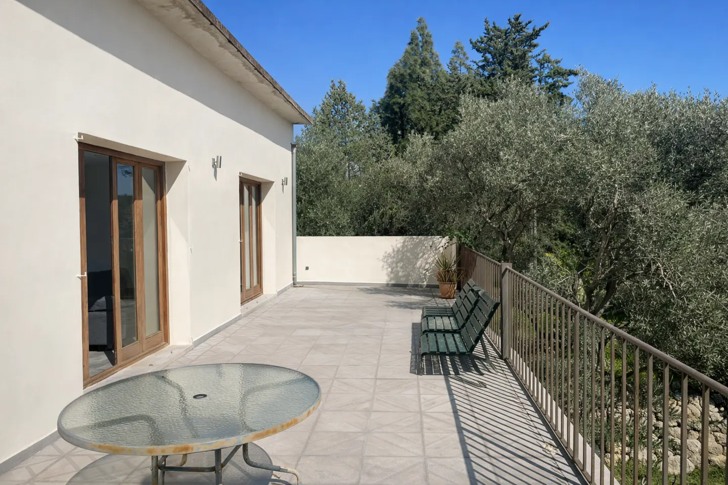 Exterior view of a house with a patio, featuring a glass table, green benches, and a metal railing overlooking trees.