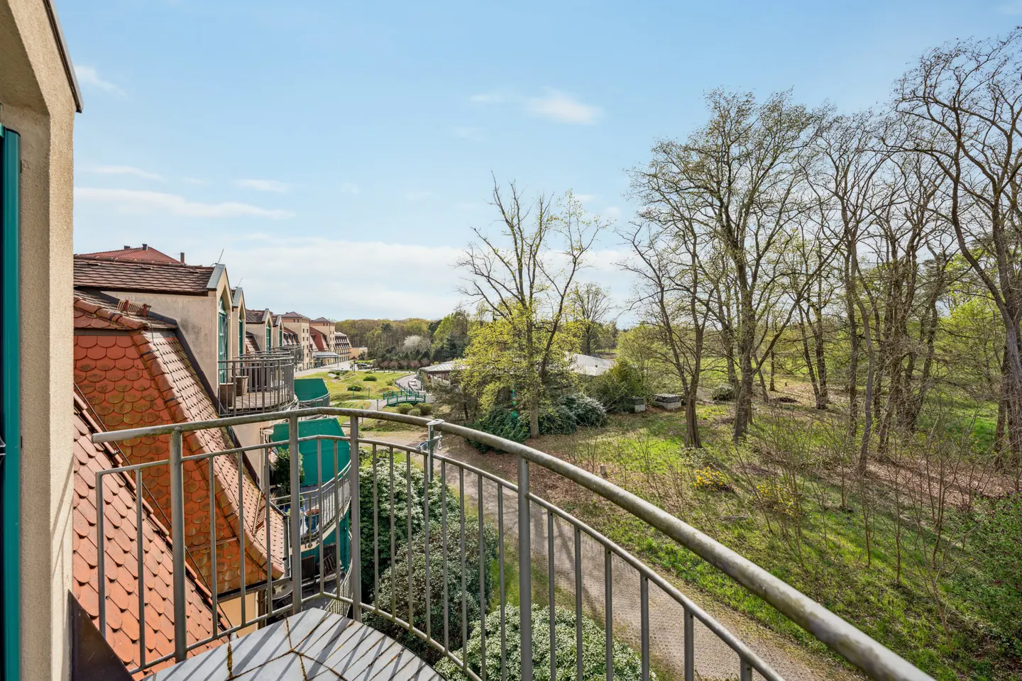 View from a balcony with metal railings overlooking a green park and buildings with red tile roofs under a blue sky.
