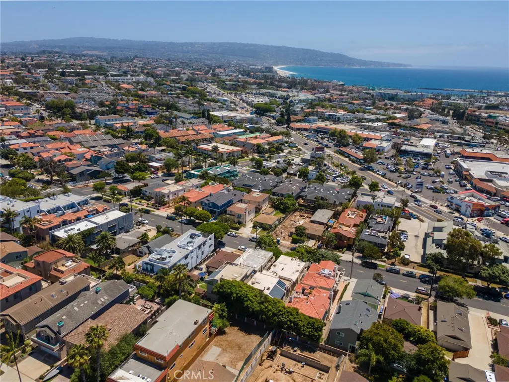 Aerial view of a coastal neighborhood with houses, palm trees, and a distant blue ocean under a clear sky.