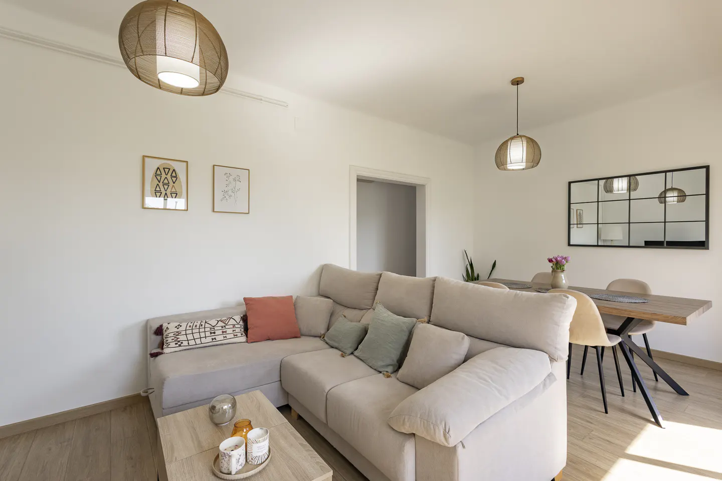 Bright living room with a beige sectional sofa, wooden coffee table, and dining table with chairs. Two pendant lights hang from the white ceiling.