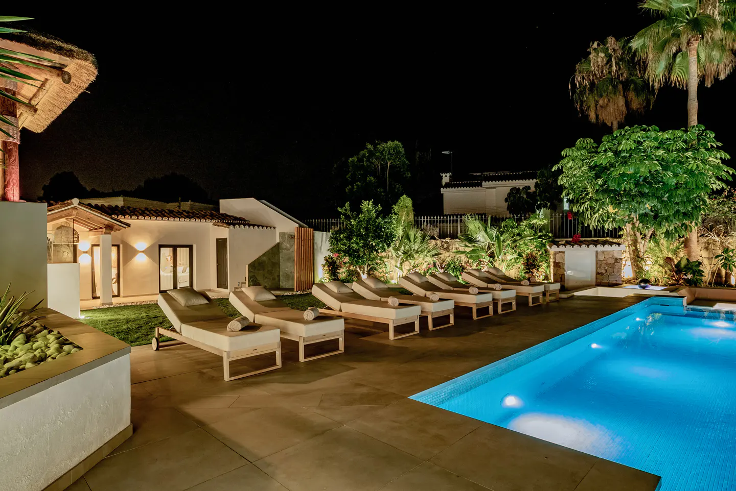 Night view of a pool with lounge chairs, white buildings, and lush greenery. The pool is lit with blue lights.