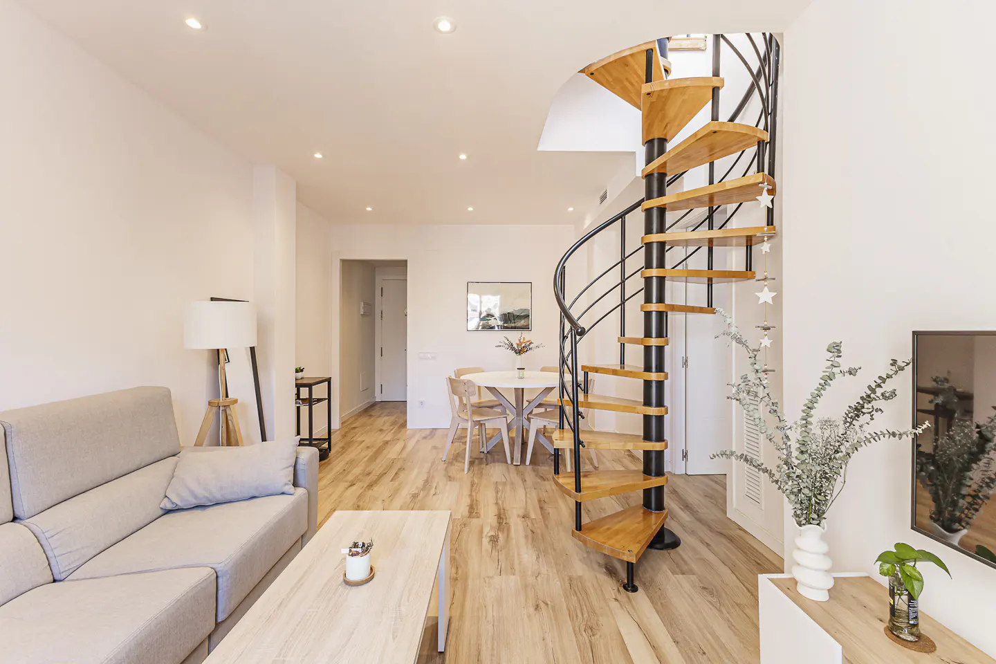 Bright living room with a gray sofa, wood floors, dining table, and a wooden spiral staircase with black metal railings.