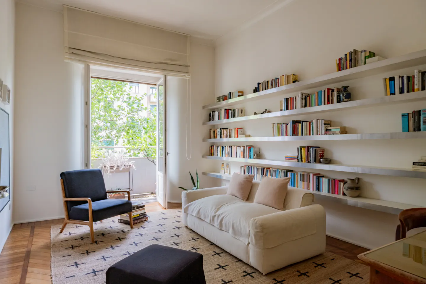 Bright living room with a white sofa, dark armchair, patterned rug, and long wall-mounted shelves filled with books.