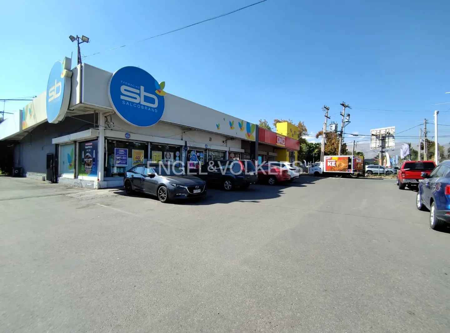 Exterior view of a strip mall with a "SB Salcobrand" pharmacy, cars parked in front, and a clear blue sky.
