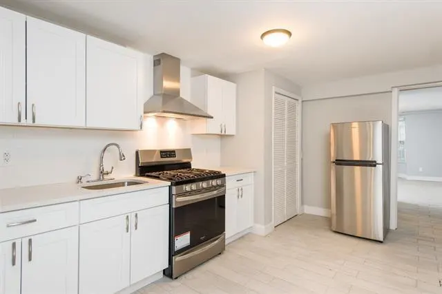 Bright kitchen with white cabinets, stainless steel appliances, and light wood-look tile flooring. A stainless steel refrigerator stands near a closet.