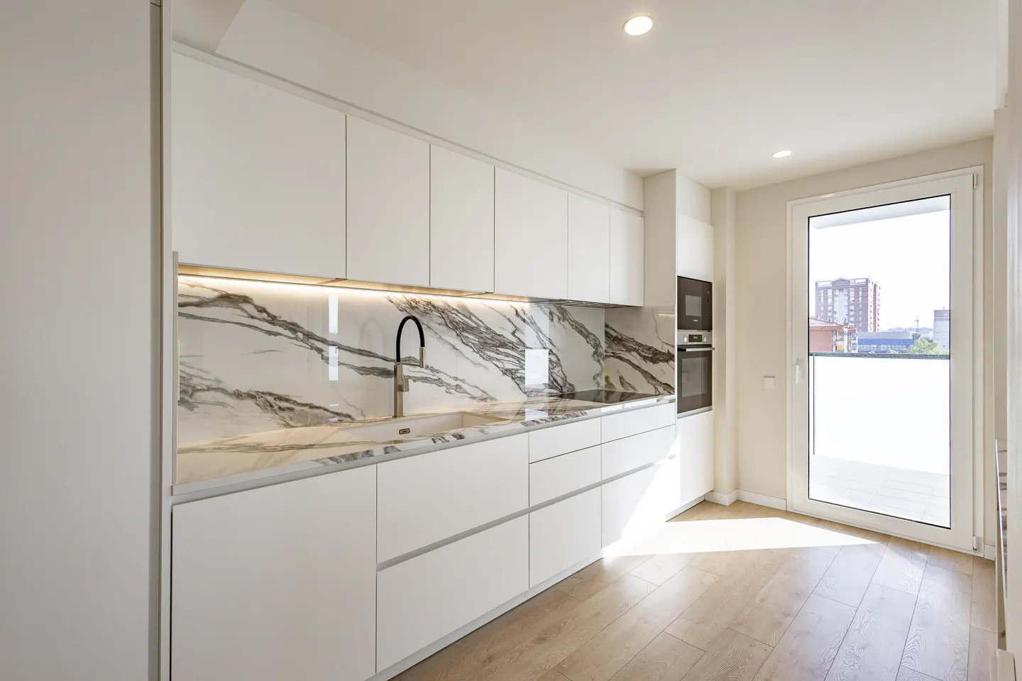 Bright, modern kitchen with white cabinets, marble backsplash, and wood floors. A black faucet is visible. A glass door leads to a balcony.