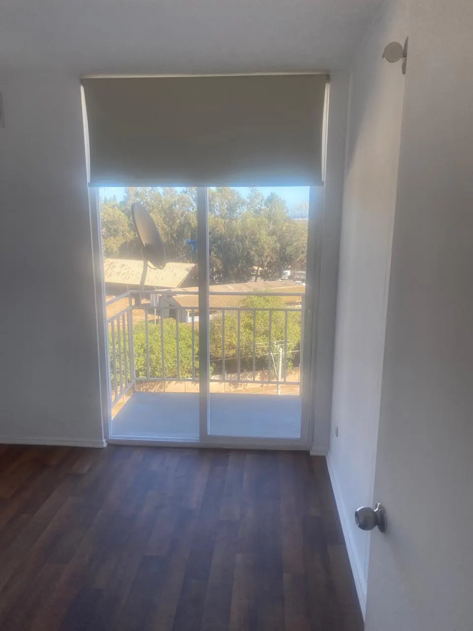 A room with dark wood floors and a sliding glass door leading to a balcony overlooking trees and a satellite dish.