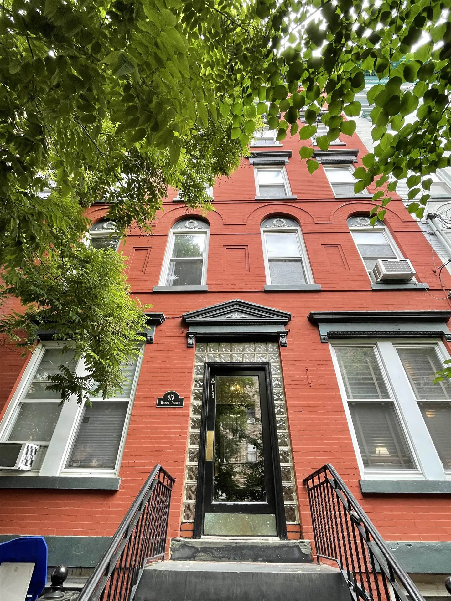 Red brick apartment building with black door and railing. Green tree branches are in the foreground.