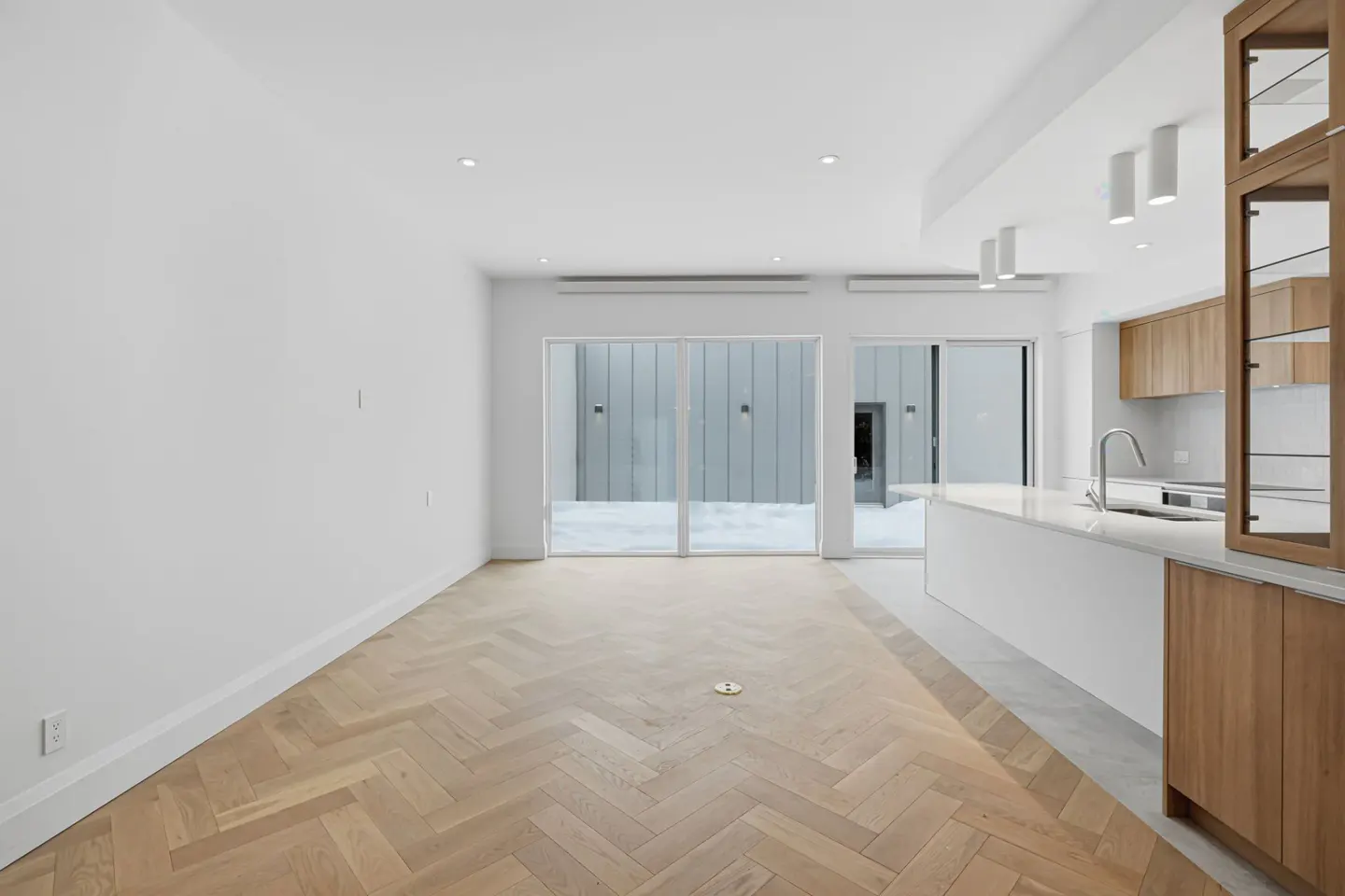 Bright, modern interior with herringbone wood floors, white walls, and sliding glass doors to a gray courtyard. Kitchen island with wood cabinets.