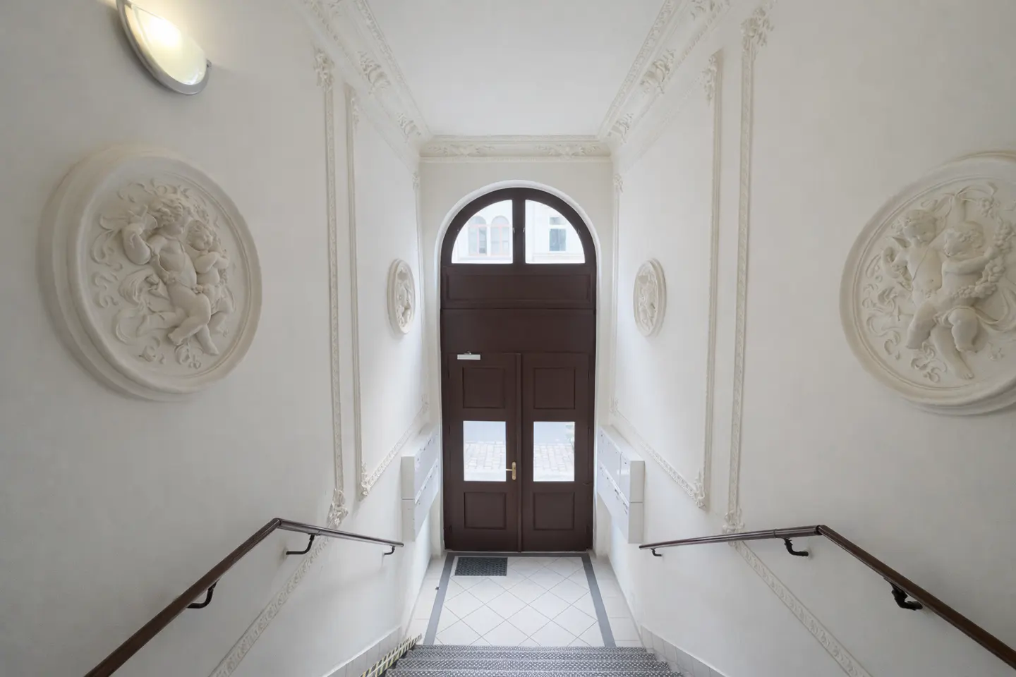 Hallway with white walls, decorative moldings, and angel reliefs. A brown arched door is at the end of the stairs with a brown handrail.