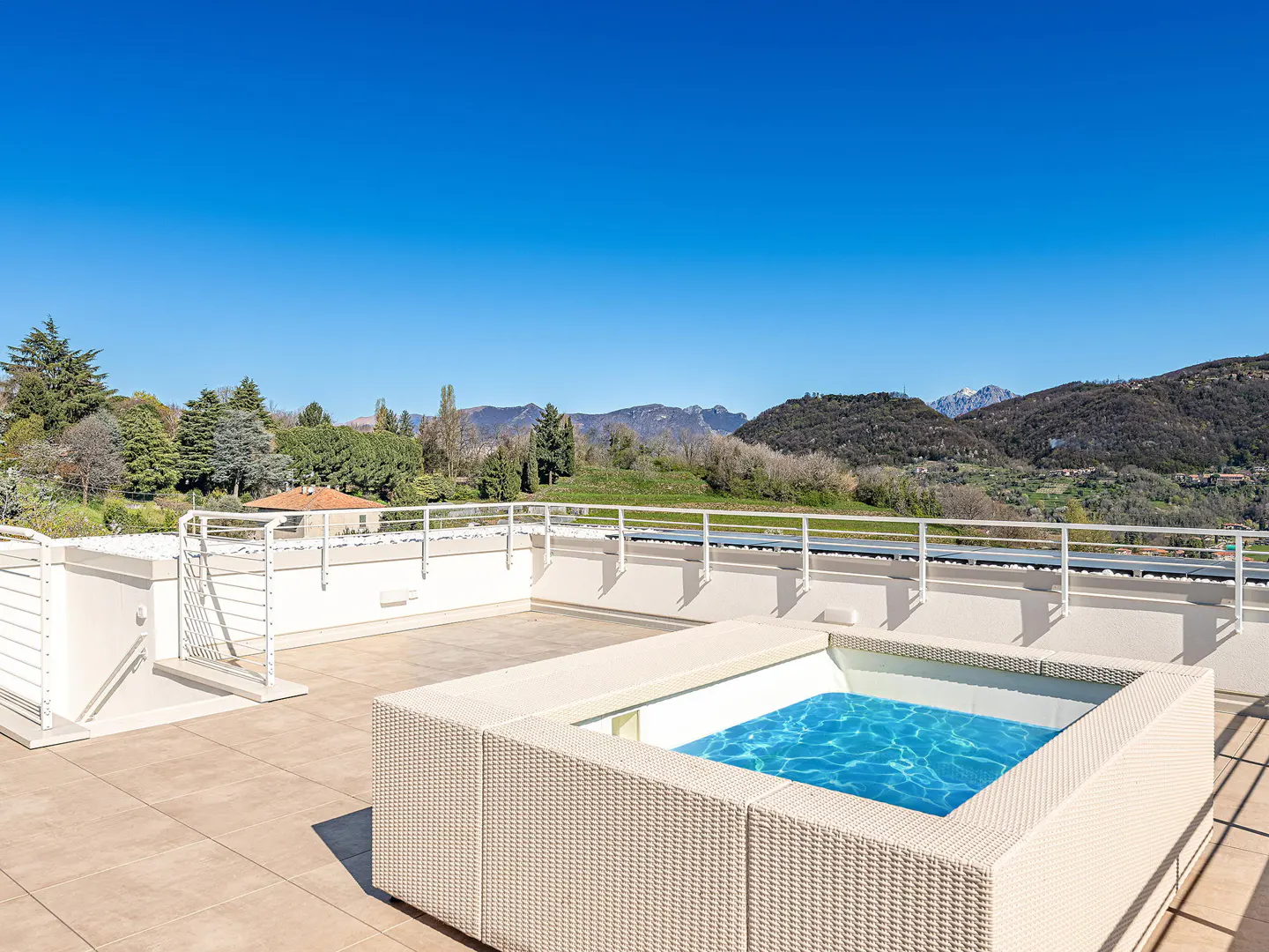 Rooftop patio with a small, square, blue pool surrounded by white wicker. Mountains and trees are in the background under a clear blue sky.