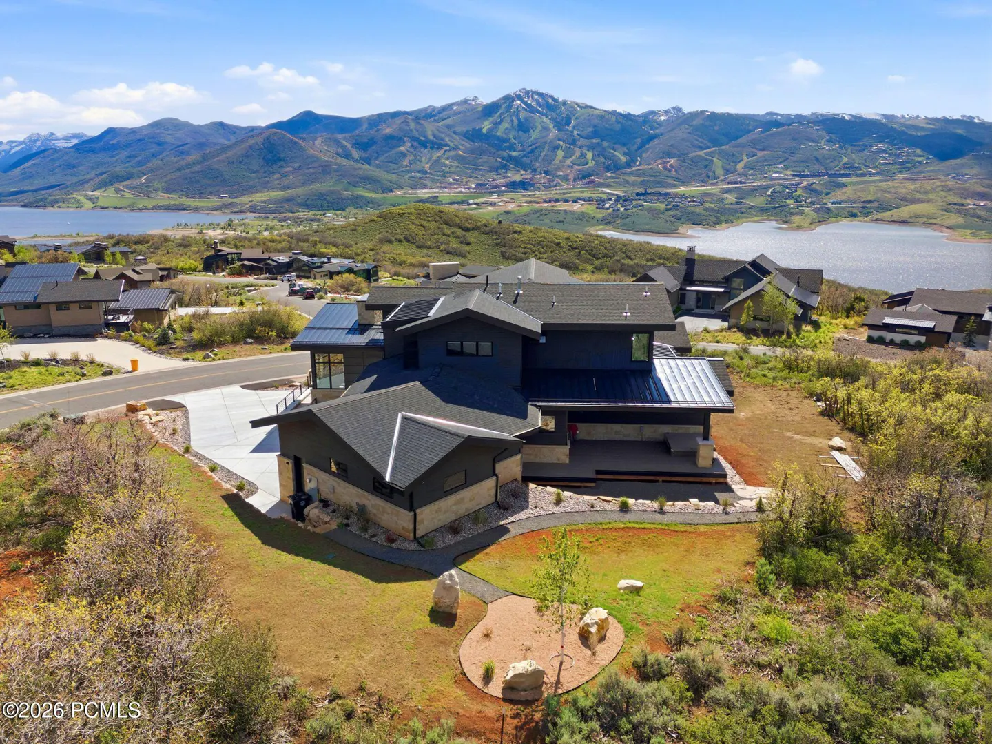 Aerial view of a modern, multi-level home with dark gray siding and black roof, set against a mountain and lake backdrop.