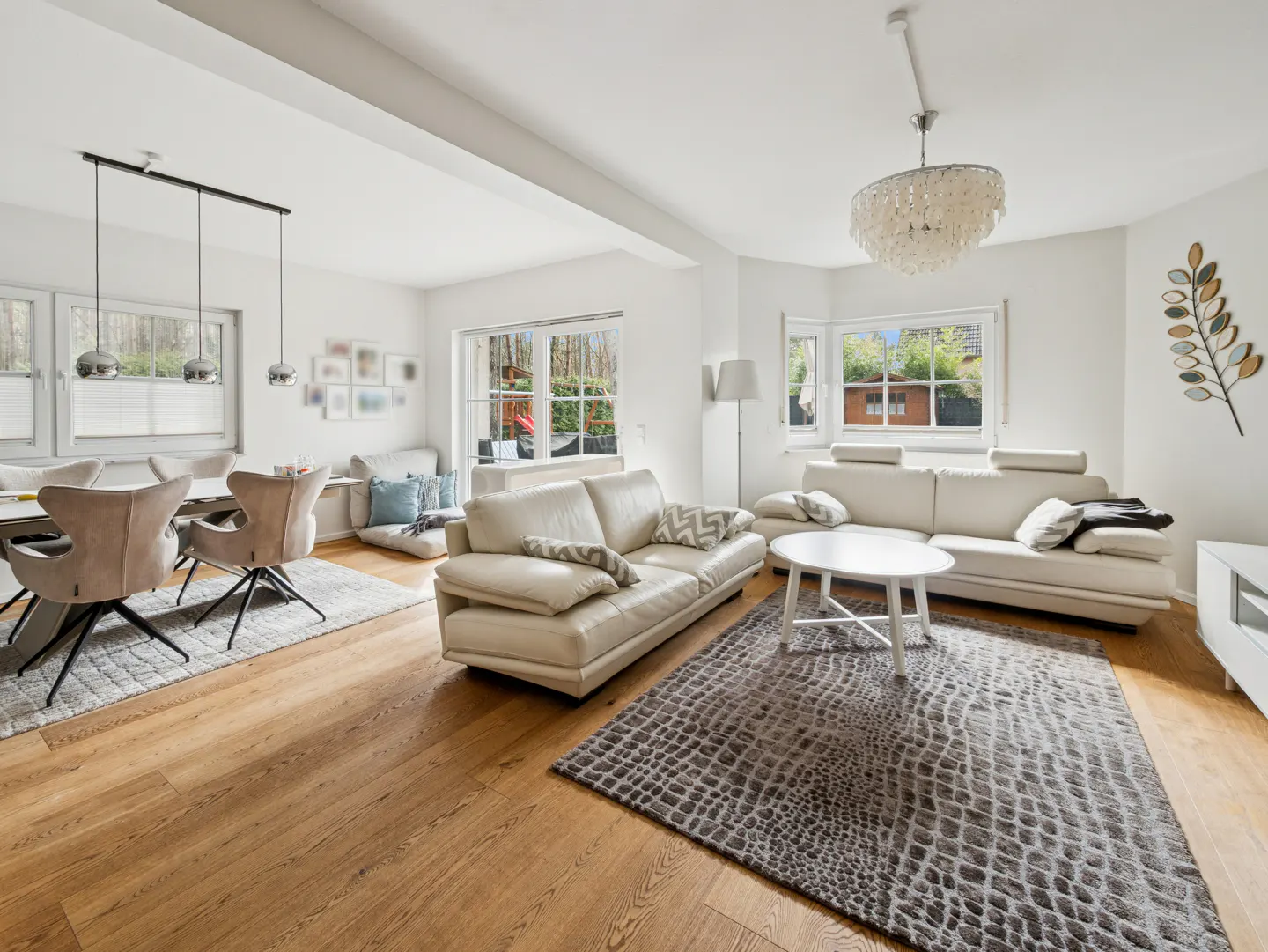Bright, open-concept living and dining area with white walls, light wood floors, and neutral furniture. Large windows offer natural light.