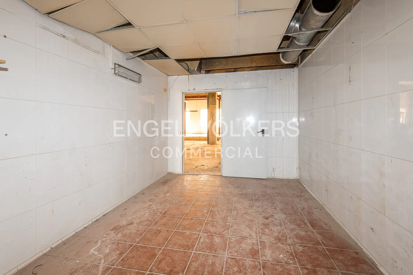 Empty commercial space with white tiled walls and brown tiled floor. A doorway leads to a brighter room. Ceiling tiles are partially missing.