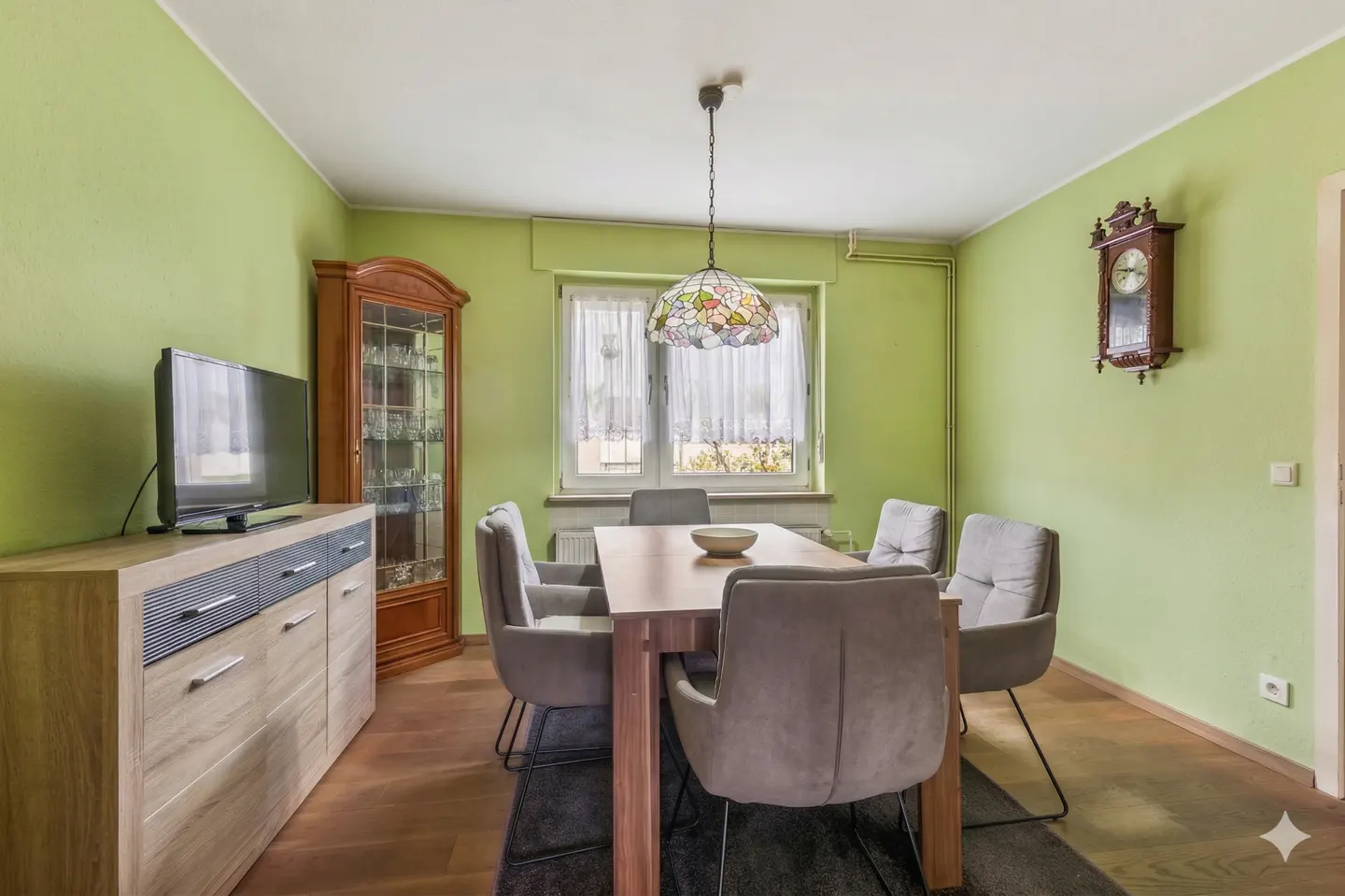 A dining room with light green walls, a wooden table with six gray chairs, and a Tiffany-style lamp. A TV and cabinet are on the left, and a clock is on the right.