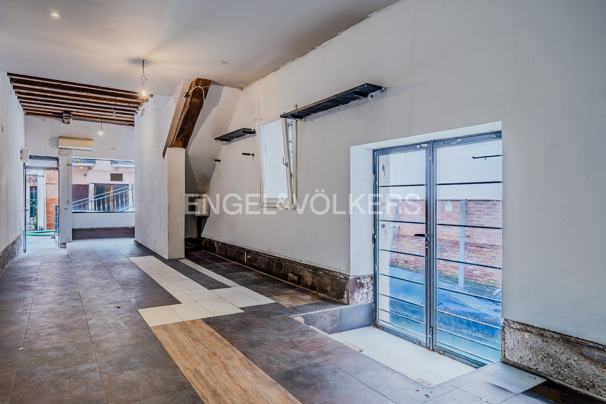 Unfurnished room with white walls, tile floor, and exposed ceiling beams. A glass door leads to an outdoor area. Engel & Völkers logo on wall.