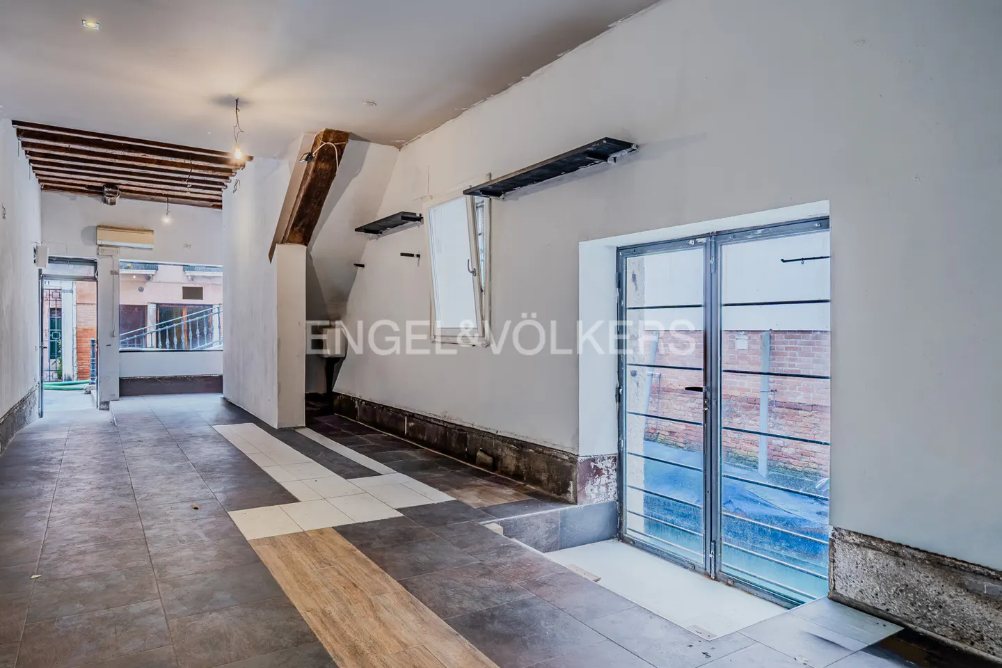 Unfurnished room with white walls, tile floor, and exposed ceiling beams. A glass door leads to an outdoor area. Engel & Völkers logo on wall.