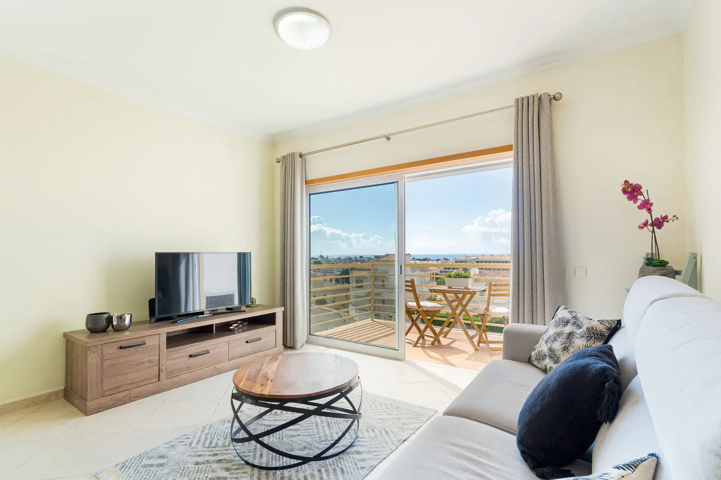 Living room with a TV, sofa, and sliding glass doors to a balcony with a table and chairs overlooking the ocean.